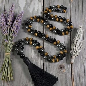 May include: A long beaded necklace featuring alternating round black beads, metallic gray beads, and golden-brown tiger eye beads, finished with a long black fabric tassel. The beads are strung in a repeating pattern along the entire length.