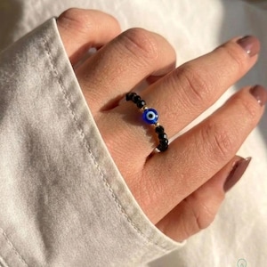 May include: A beaded ring featuring small, faceted black beads and a central blue, white, and black evil eye charm. The ring is accented with tiny gold-toned beads placed on either side of the evil eye charm, creating a circular band design.