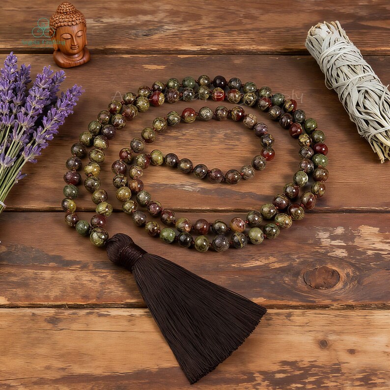 May include: A beaded mala necklace with a dark brown tassel. The necklace features green, brown, and red beads. A small wooden Buddha statue, a bundle of lavender, and a sage bundle are also in the image.