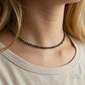 May include: A beaded necklace with small, faceted beads in shades of green, red, and gray. The necklace is worn around the neck and appears to be made of natural gemstones. The beads are strung together to create a delicate choker style.