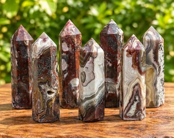 May include: Seven polished stone towers arranged in a group, each featuring unique swirling patterns of red, brown, white, and gray. The hexagonal columns have pointed tops and are displayed on a textured wooden surface with a blurred green background.