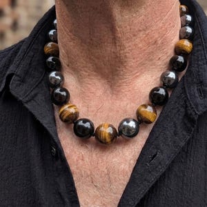 May include: A beaded necklace featuring alternating large spherical beads in tiger's eye, polished black onyx, and metallic hematite. The beads are strung together in a repeating pattern, creating a substantial circular design worn around the neck.