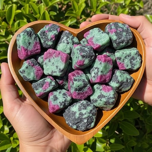 May include: A heart-shaped wooden bowl filled with rough-cut ruby zoisite stones. The stones display a mix of green, pink, and black colors. The bowl is held by two hands against a backdrop of green foliage.