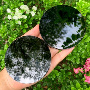 May include: Two circular, polished black obsidian stones with smooth, reflective surfaces. Each stone is perfectly round and dark, showing clear reflections of the surrounding environment on their glossy, mirror-like faces.