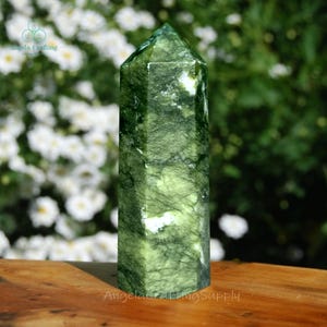 May include: A polished, hexagonal green crystal tower featuring deep forest green hues, intricate black veining, and scattered white inclusions. The stone has a smooth, reflective surface with a pointed top, showcasing natural mineral patterns.