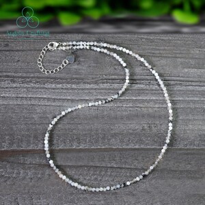 May include: Delicate necklace featuring small, round, white and gray beads. The necklace has a silver-colored chain and clasp. The beads are strung together to create a simple, elegant design. The necklace is displayed on a gray wooden surface.