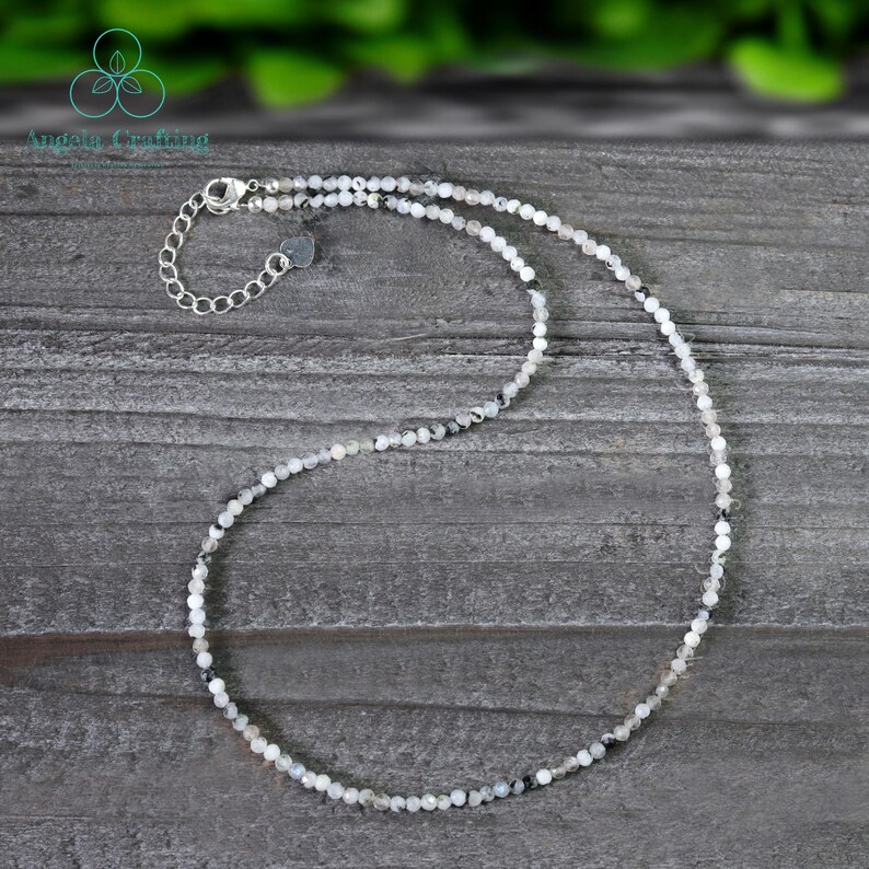 May include: Delicate necklace featuring small, round, white and gray beads. The necklace has a silver-colored chain and clasp. The beads are strung together to create a simple, elegant design. The necklace is displayed on a gray wooden surface.
