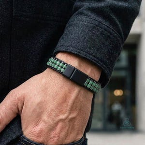 Pode incluir: Uma pulseira com duas filas paralelas de pequenas contas quadradas verde-sálvia, separadas por um fino divisor de metal escuro. A pulseira é fixada com um fecho retangular de metal preto fosco que fica visível no pulso.