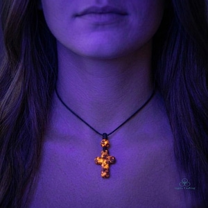 May include: A cross-shaped pendant with a glowing orange and yellow mottled pattern suspended on a thin black cord. The pendant features a small spherical bead directly above the cross, both emitting a bright light against the dark background.