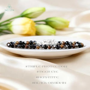 May include: A beaded bracelet with black, brown, and silver beads rests on a white plate. The bracelet is made of tiger eye, hematite, and black obsidian. The background features yellow and green flowers.