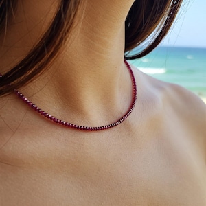 May include: A delicate necklace made of small, dark red gemstones. The necklace is worn by a person with long brown hair, and the background is a blurry image of a beach.