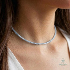 May include: A delicate, light blue beaded necklace. The necklace is composed of small, round beads, creating a subtle and elegant accessory. The necklace is worn around the neck, showcasing its simple design and color.