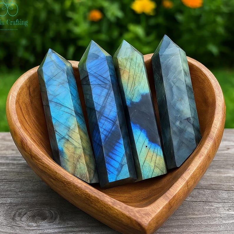 May include: Four polished labradorite crystal towers with blue, green, and gold iridescence are displayed in a heart-shaped wooden bowl. The crystals have a faceted, pointed top and are arranged side by side. The bowl is made of a light brown wood.