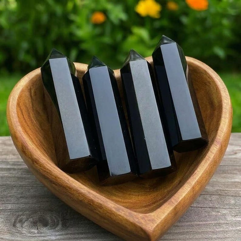 May include: Four polished black obsidian crystal points with hexagonal facets arranged upright inside a smooth, heart-shaped wooden bowl. The dark, reflective surfaces of the stone towers contrast with the natural light brown wood grain.