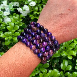 May include: A multi-strand bracelet composed of polished, spherical purple and dark violet beads. The beads exhibit natural variations in color and tone, creating a deep, rich appearance. The bracelet is arranged in several rows of smooth, round stones.