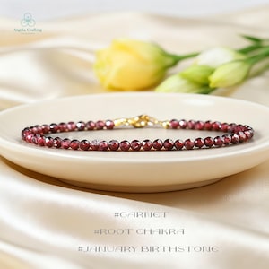 May include: A delicate bracelet composed of small, faceted, deep red garnet beads arranged in a continuous circular strand. The bracelet features a gold-toned metal lobster claw clasp and jump ring closure, providing a secure and functional connection.