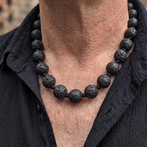 May include: A necklace composed of large, round, black lava stone beads with a porous, textured surface. The beads are strung together on a dark cord, creating a uniform circular design that rests around the neck over a black collared shirt.
