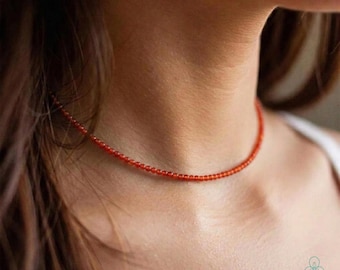 May include: A delicate, red beaded necklace. The necklace is a choker style, composed of small, round beads. The beads are a vibrant, warm red color. The necklace is worn around the neck.