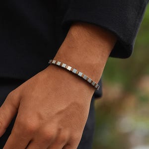Op de afbeelding: Een armband met een herhalend patroon van vierkante kralen in afwisselende zilver- en bruintinten. De armband wordt om een pols gedragen, met een zwarte mouw op de achtergrond. De armband heeft een eenvoudig, modern ontwerp.