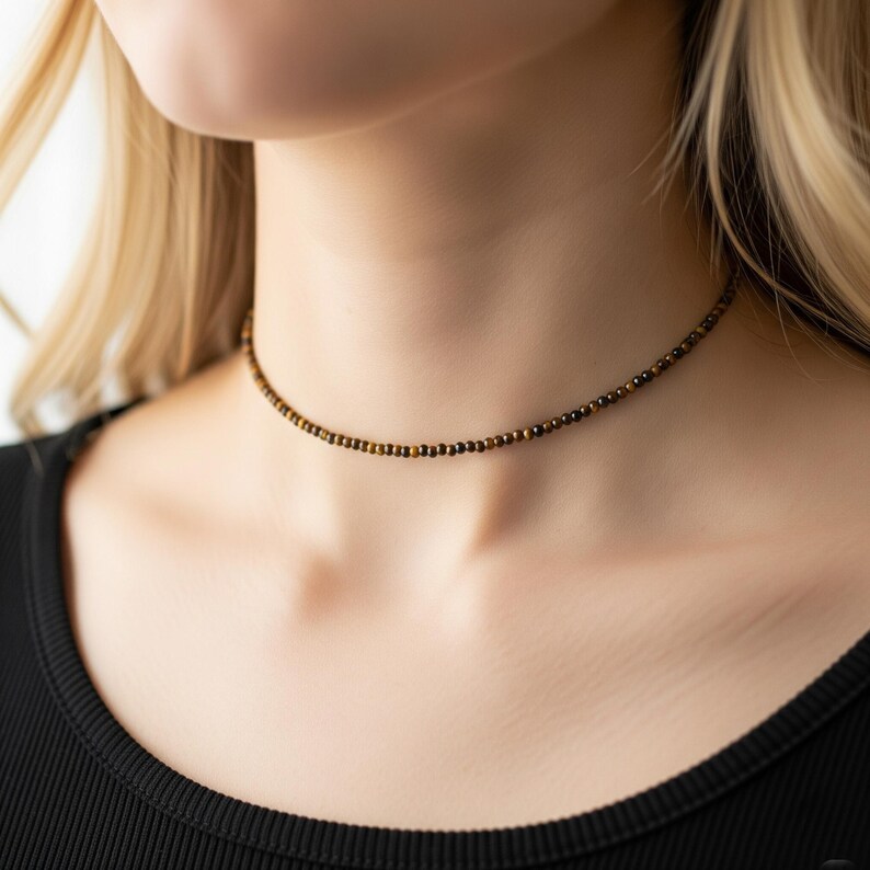 May include: A delicate choker necklace featuring small, round beads in shades of brown and gold. The necklace is worn around the neck, resting just above the collarbone. The beads create a subtle, elegant accessory.