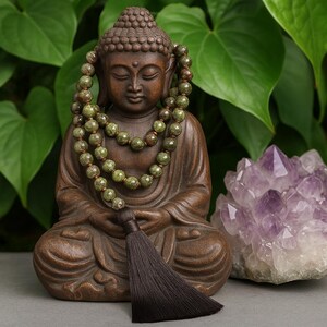 May include: A brown Buddha statue with a green beaded necklace and a dark brown tassel. A large purple crystal is to the right of the statue. The background features green leaves.