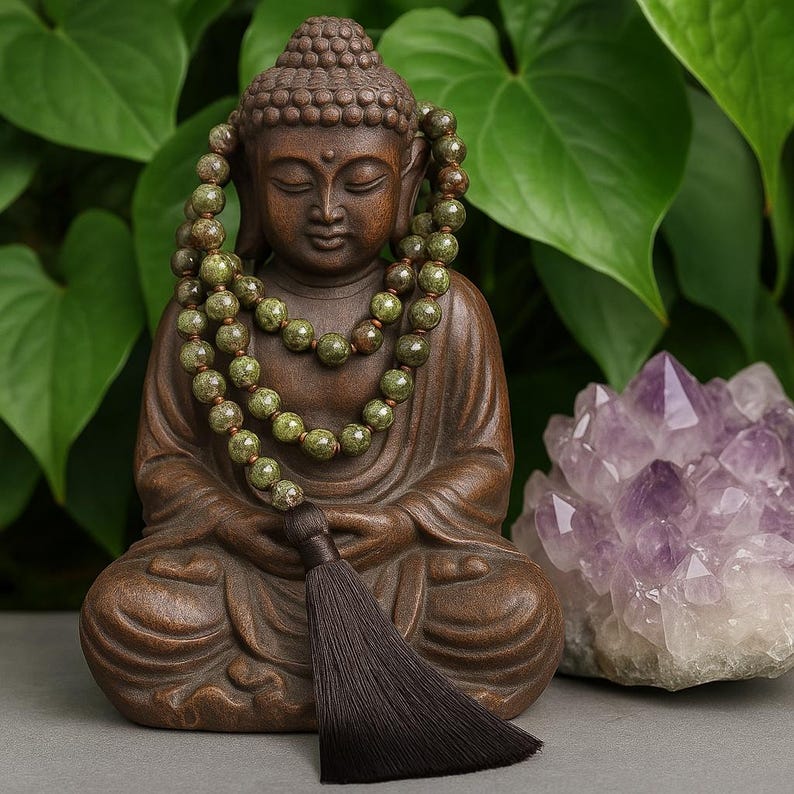 May include: A brown Buddha statue with a green beaded necklace and a dark brown tassel. A large purple crystal is to the right of the statue. The background features green leaves.