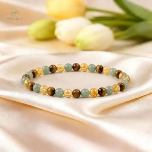Op de afbeelding: Een armband met ronde edelstenen in bruin, geel en groen. De kralen zijn geregen en vormen een cirkel. De armband ligt op een witte satijnen achtergrond, met onscherpe bloemen. Een sieraad.