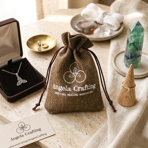 May include: A brown burlap drawstring pouch with the white text Angela Crafting Spiritual Healing Meditation, a silver necklace in a dark box, a gold ring on a wooden cone, a green crystal point, and a business card with the same logo.