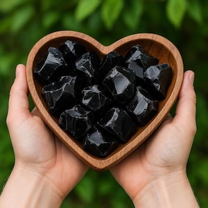 May include: A heart-shaped wooden bowl filled with multiple raw, glossy black obsidian stones. The dark, reflective surfaces of the stones contrast with the natural brown wood grain of the bowl, which features smooth, curved edges and a deep interior.