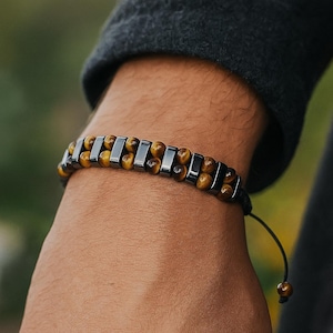 May include: A beaded bracelet featuring alternating tiger's eye and dark gray rectangular beads. The bracelet is strung on a black cord with an adjustable closure. The bracelet is a stylish accessory.