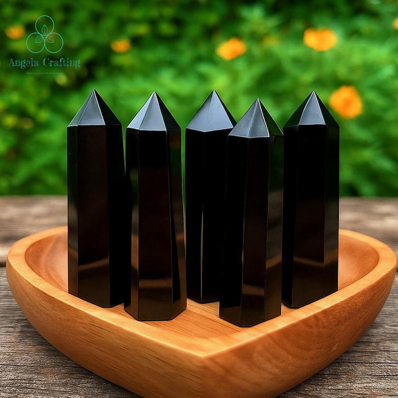 May include: Five polished black obsidian crystal points with hexagonal bases and pointed tops, arranged in a vertical formation inside a smooth, light-colored wooden bowl shaped like a heart, featuring a reflective surface on each stone.