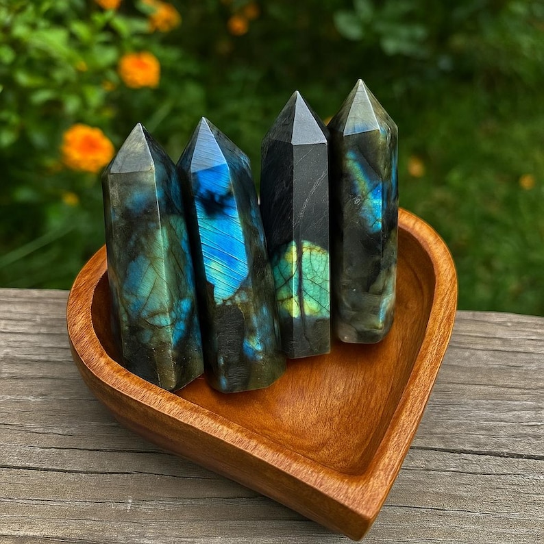 May include: Four polished labradorite crystal towers with blue, green, and gray iridescent flashes are displayed in a heart-shaped wooden bowl. The crystals have a pointed top and are arranged in a row. The bowl is made of light brown wood.