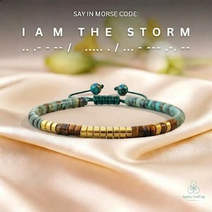 Könnte beinhalten: Ein Armband mit türkisfarbenen, braunen und goldfarbenen Perlen. Der Text "I AM THE STORM" steht in Morsecode über dem Armband. Das Armband liegt auf einer cremefarbenen Satin-Oberfläche.