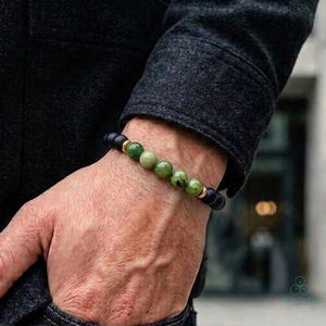 May include: A beaded bracelet featuring a combination of black, green, and gold beads. The bracelet is composed of matte black beads, interspersed with green and gold-colored beads. The bracelet is worn on a wrist.