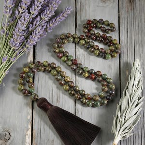 May include: A collection of beaded necklaces with a brown tassel, alongside a bundle of lavender and a sprig of sage. The necklaces feature green, brown, and red beads. The items are arranged on a weathered, gray wooden surface.