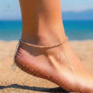 Peut inclure: Un bracelet de cheville à perles avec un charm en forme de cœur. Le bracelet de cheville est orné de petites perles orange clair et d'une chaîne argentée avec un pendentif en forme de cœur. L'image est prise sur une plage de sable avec l'océan en arrière-plan.