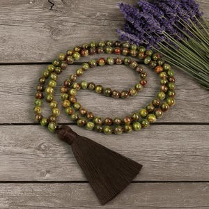 May include: A beaded necklace with green and brown beads and a brown tassel. The necklace is draped on a wooden surface next to a bundle of purple lavender.