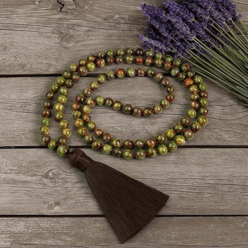 May include: A beaded necklace with green and brown beads and a brown tassel. The necklace is draped on a wooden surface next to a bundle of purple lavender.