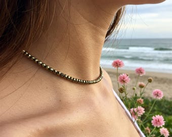 May include: A gold choker necklace made of small, faceted beads. The necklace is worn by a person with long brown hair, and the background is a blurry image of a beach with ocean waves and pink flowers.