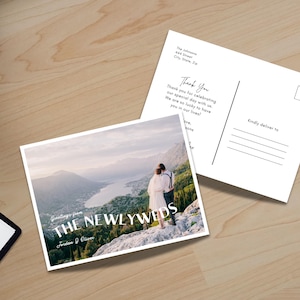May include: Two white postcards on a wooden surface. One postcard features a scenic photo of a couple on a mountain with the text "Greetings from THE NEWLYWEDS". The other postcard has a "Thank You" message and address lines.