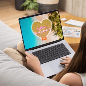 Puede incluir: Un port&aacute;til plateado que muestra una interfaz de juego con el texto "Callie's island jeopardy" dentro de un coraz&oacute;n dorado. La pantalla muestra una escena de playa con agua azul y palmeras. El juego tiene un bot&oacute;n "Start".