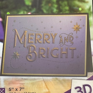 May include: A 3D embossing folder for crafting, featuring the text "Merry and Bright" in gold lettering on a purple background with gold stars. The folder measures 5 inches by 7 inches (12.7 cm by 17.8 cm).