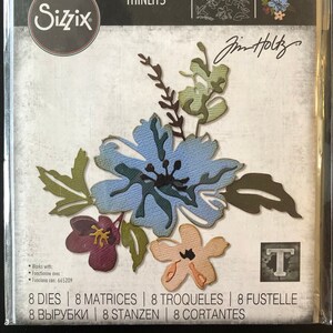 May include: A set of 8 metal dies for crafting featuring a floral design with blue, green, pink, and purple flowers. The dies are designed by Jim Holtz and are part of the Sizzix Thinlits collection. The package includes the text "8 DIES | 8 MATRICES | 8 TROQUELES | 8 FUSTELLE | 8 ВЫРУБКИ | 8 STANZEN | 8 CORTANTES".