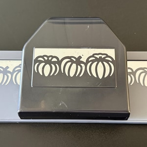 May include: A dark gray paper punch tool featuring a wide rectangular base and a central handle. The device is designed to create a repeating border pattern of three distinct pumpkin shapes, visible as cutouts on the front of the plastic casing.