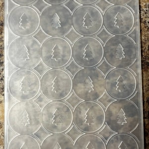 May include: A clear plastic sheet with a repeating pattern of 20 circular cutouts, each containing a silhouette of a Christmas tree. The sheet is labeled "Cutterbug" and "Provo Craft".
