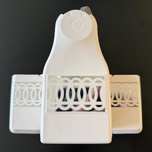 May include: A white paper craft punch with a decorative oval design. The punch has a lever on top and two side compartments with the same design. The punch is designed for creating decorative edges on paper or cardstock.