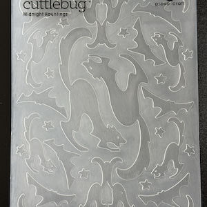 May include: A clear plastic Cricut Cuttlebug embossing folder labeled Midnight Hauntings by Provo Craft. The rectangular plate features recessed and raised patterns of cats, bats, stars, and crescent moons across the entire surface for paper crafting.