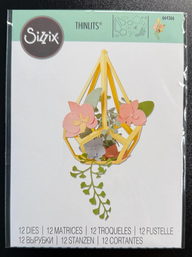Puede incluir: Un juego de 12 troqueles Sizzix Thinlits 664366. El paquete muestra una estructura de terrario geom&eacute;trica amarilla llena de orqu&iacute;deas rosas, hojas verdes y follaje colgante hechos de papel, todo presentado en una funda de pl&aacute;stico transparente.