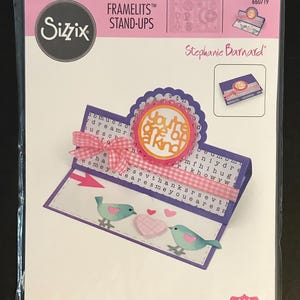 May include: Sizzix Framelits Stand-Ups die set by Stephanie Barnard. The package features a sample card with a pink ribbon, two teal birds, and the phrase "You're one of a kind". Includes 19 dies.