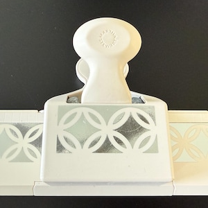 May include: A white Martha Stewart craft punch with a geometric design in silver and light green. The punch has a large handle and a decorative pattern on the front and sides, ideal for paper crafting.